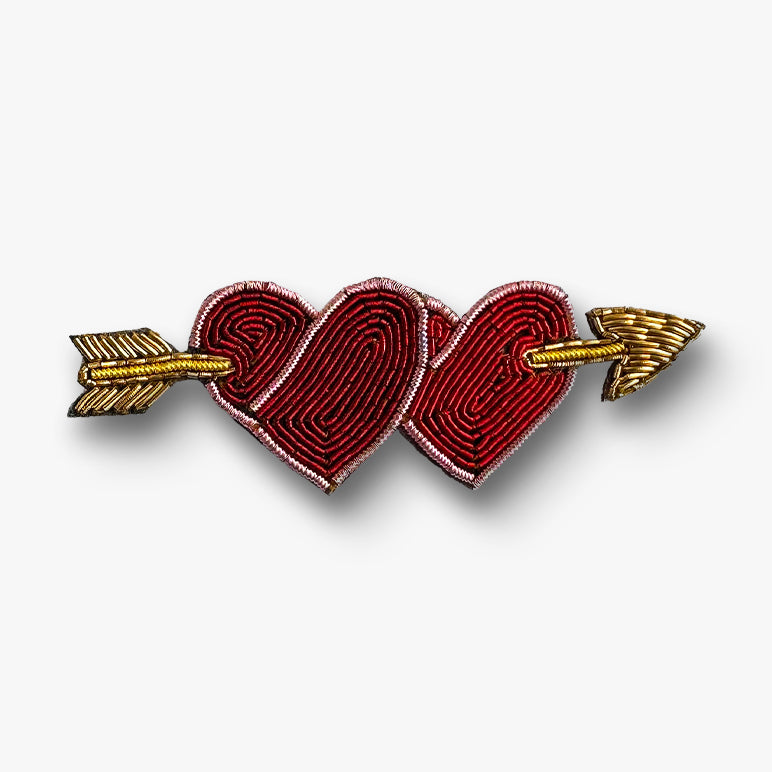 Embroidered brooch pin of two interlocking hearts with gold arrows on a white background