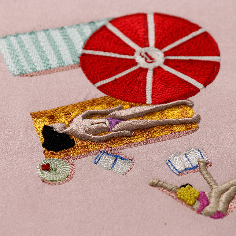Embroidered scene of a person sunbathing under a red umbrella on a pink background
