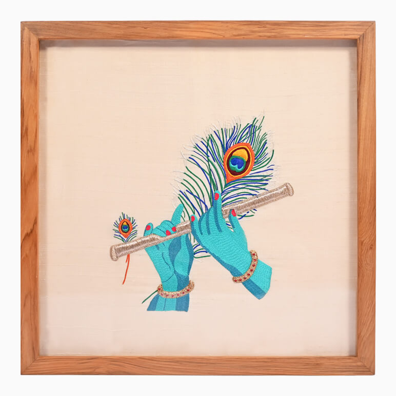 Embroidery of hands playing a flute with peacock feathers, framed in wood.