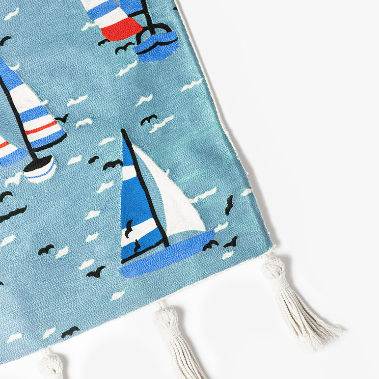 Corner detail of A Summer Sail sailboat wall hanging—hand-embroidered blue sea with striped sails, white wave stitches, black birds, and tasselled edge.