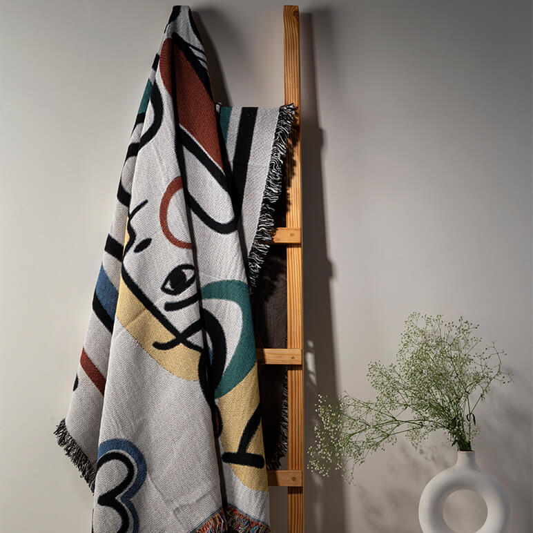 Decorative blanket with abstract patterns hanging on a wooden ladder against a plain wall.