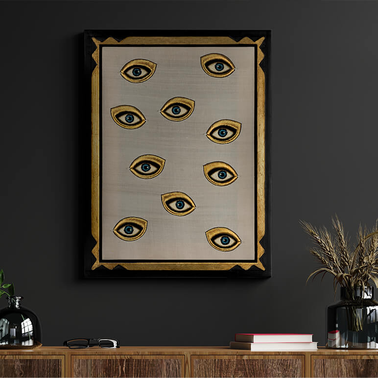 Framed artwork with eye designs on a dark wall above a wooden shelf.