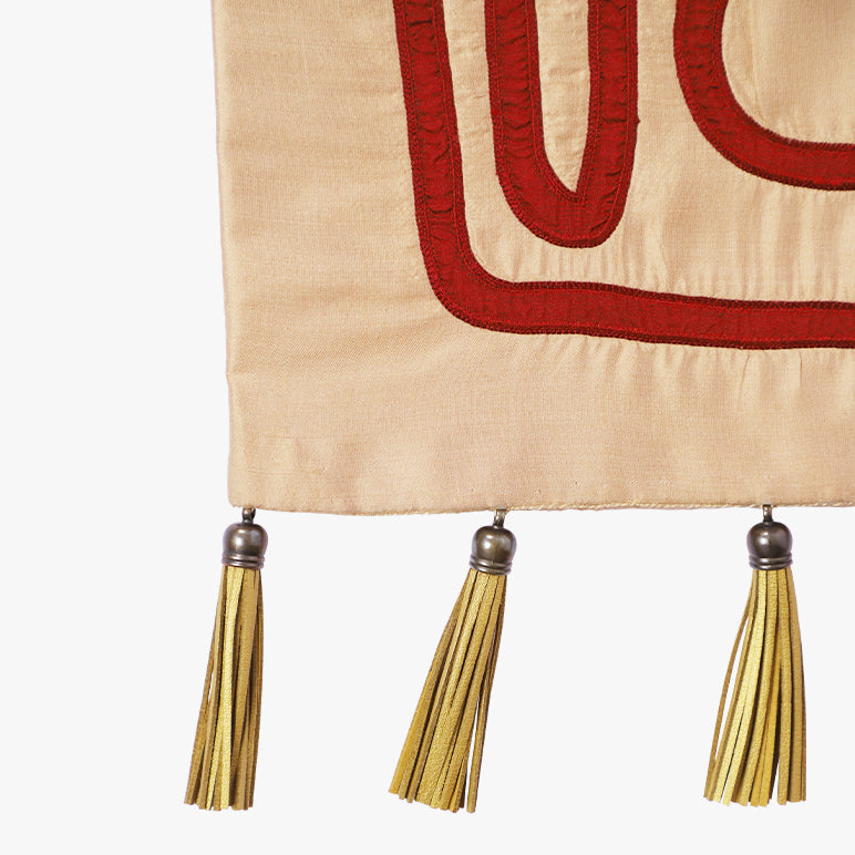 Close-up of the Crimson Whispers tapestry—crimson embroidered lines on warm beige fabric with gold tassels and metal caps along the bottom edge.