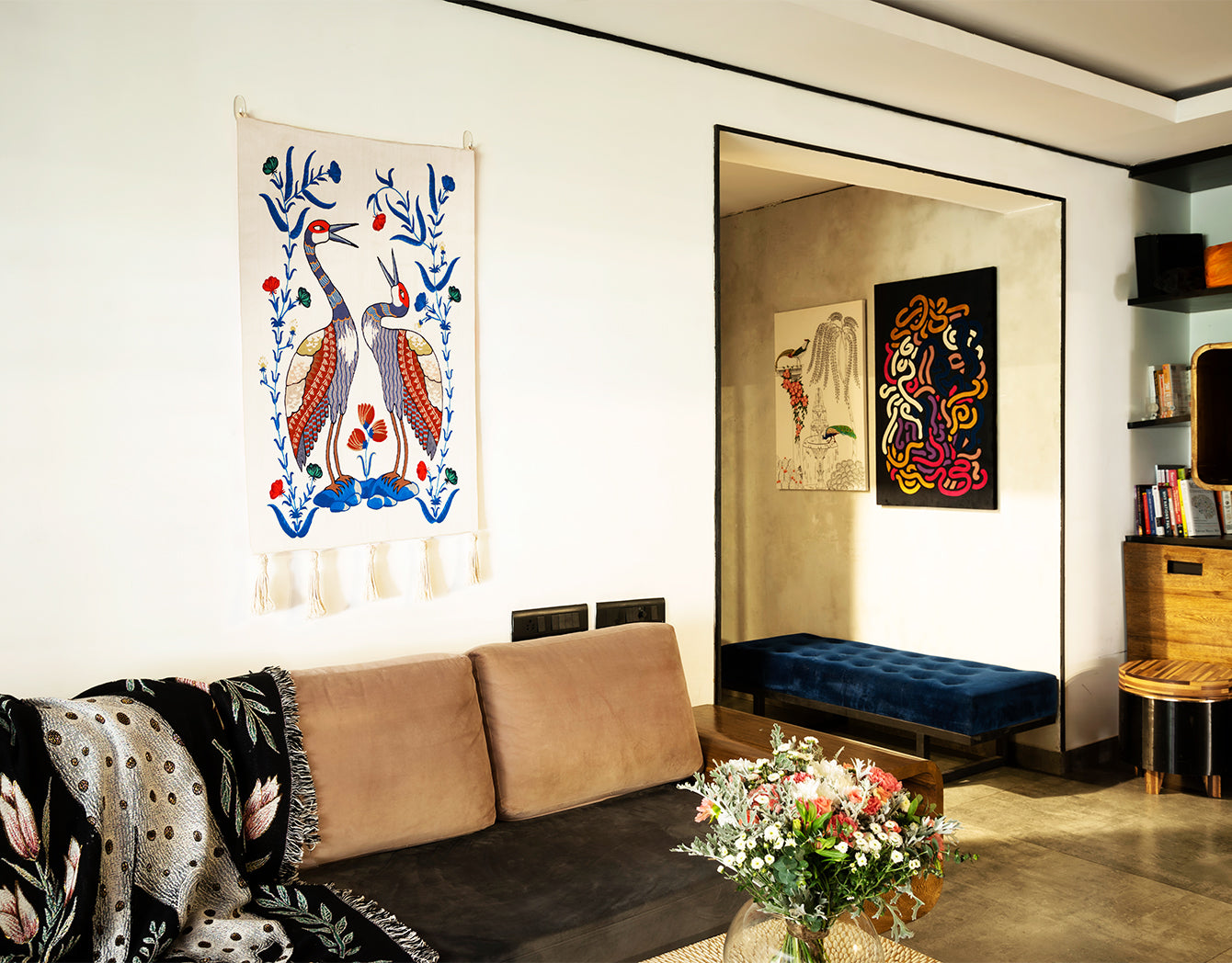 Songs of the Crane embroidered wall hanging—two stylized cranes with blue floral vines on an ivory textile with tassels—displayed as modern wall decor above a tan sofa in a bright living room.