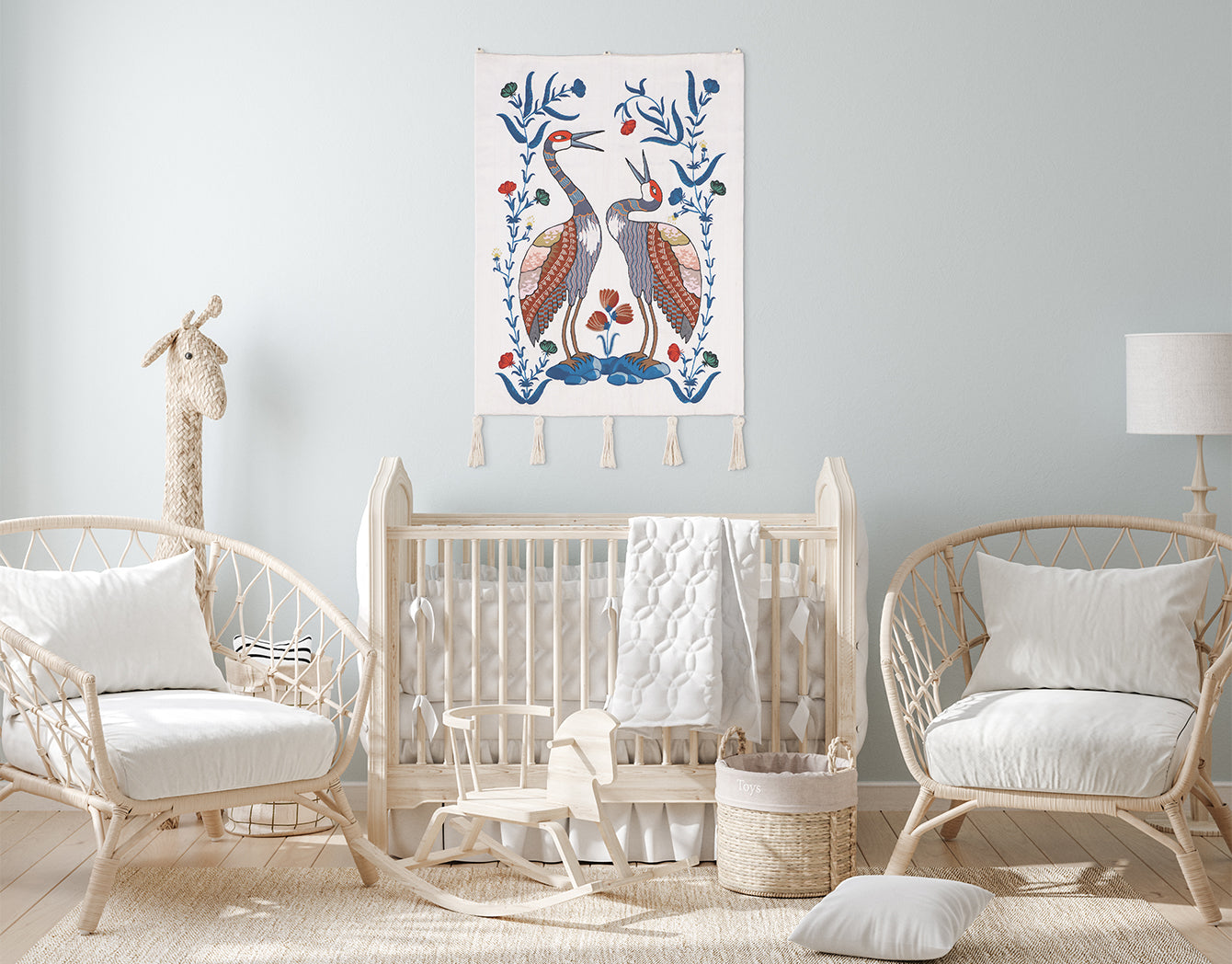 *Songs of the Crane* embroidered wall hanging—two stylized cranes with blue floral vines on ivory—hung above a natural-wood crib in a light, airy nursery.
