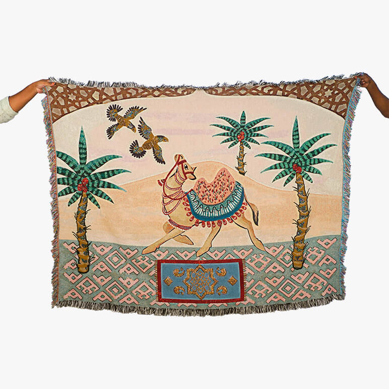 Designer throw blanket with a camel and palm tree design held by two hands.