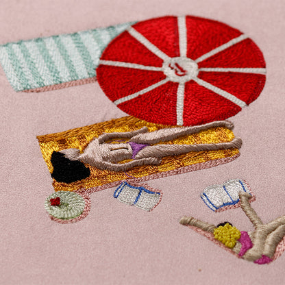 Embroidered scene of a person sunbathing under a red umbrella on a pink background