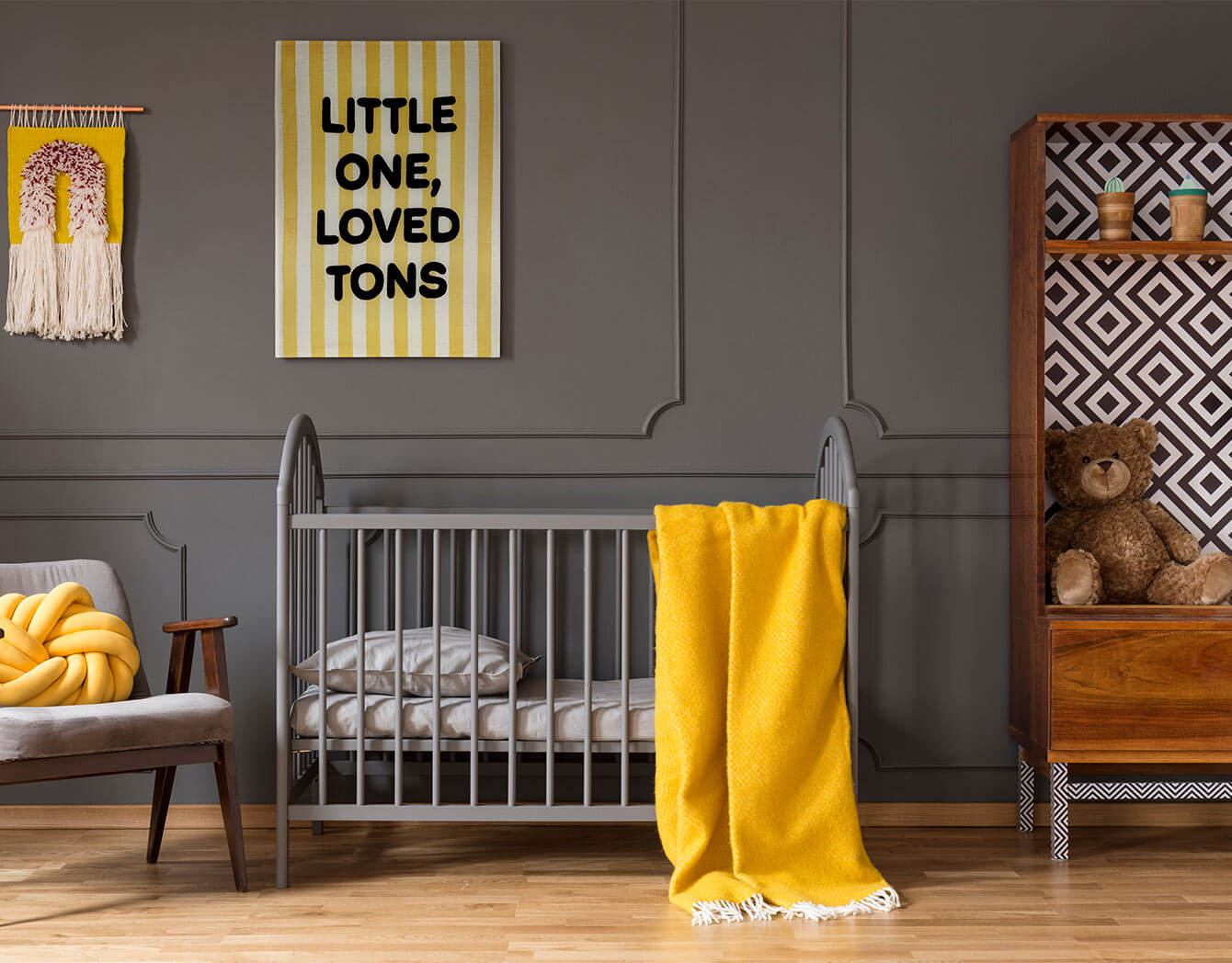 Nursery room with gray crib, yellow blanket, and teddy bear against a gray wall with a striped gender-neutral wall art.