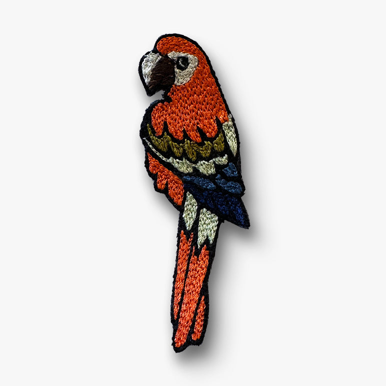Silk-thread parrot brooch pin in orange body with navy blue wings and olive accents on a white background