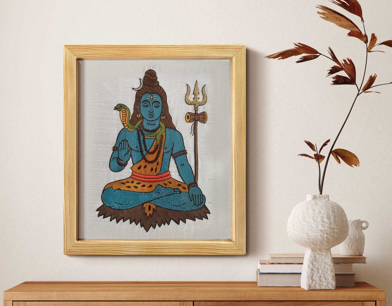 Framed artwork of a seated Lord Shiva with a trident on a wall above a wooden shelf.