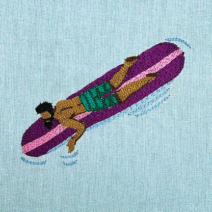 Embroidery of a person on a purple surfboard with green shorts on a light blue fabric background