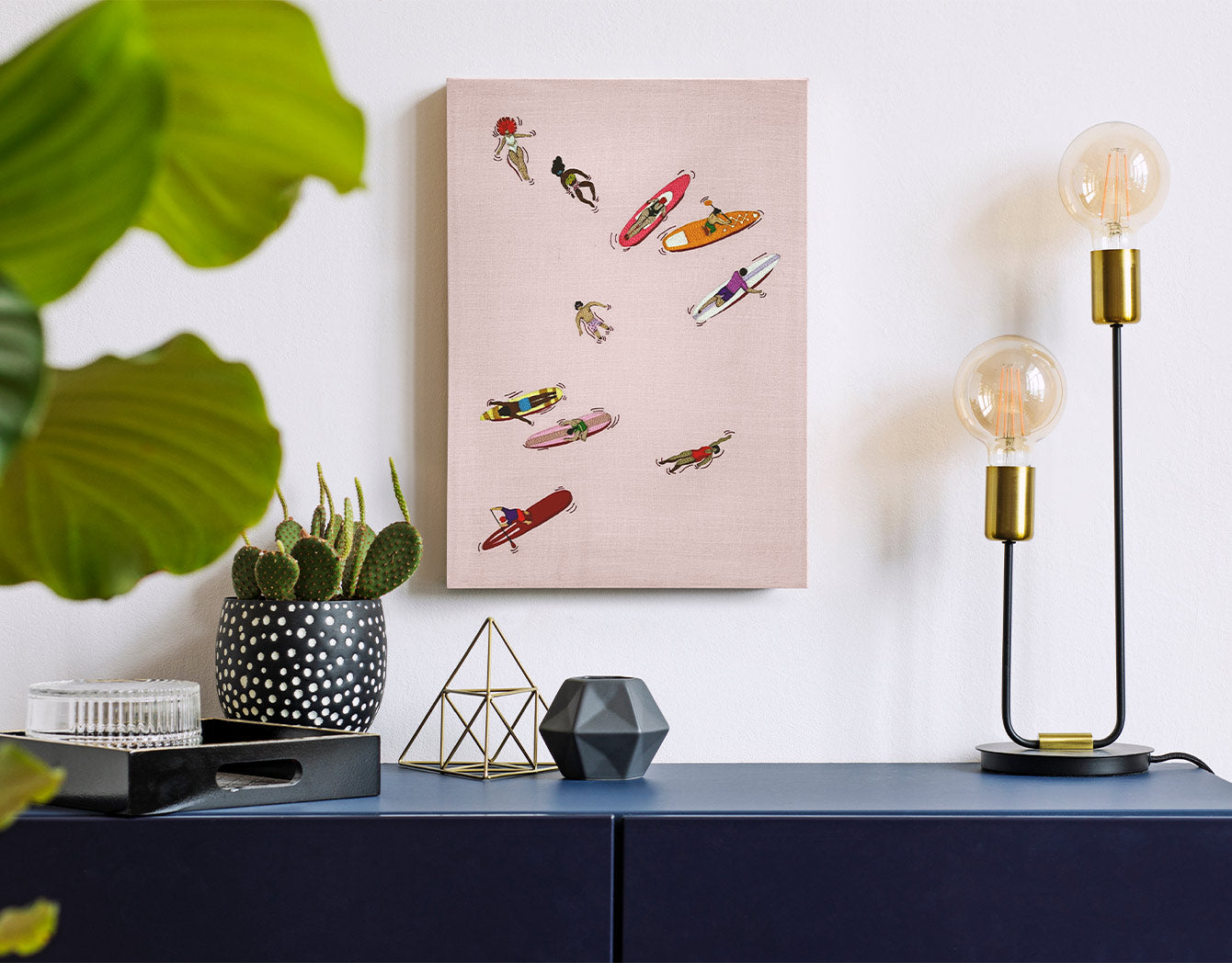 Decorative modern beach wall art with colorful embroidery on a pink background, placed above a blue cabinet with a lamp and plant.