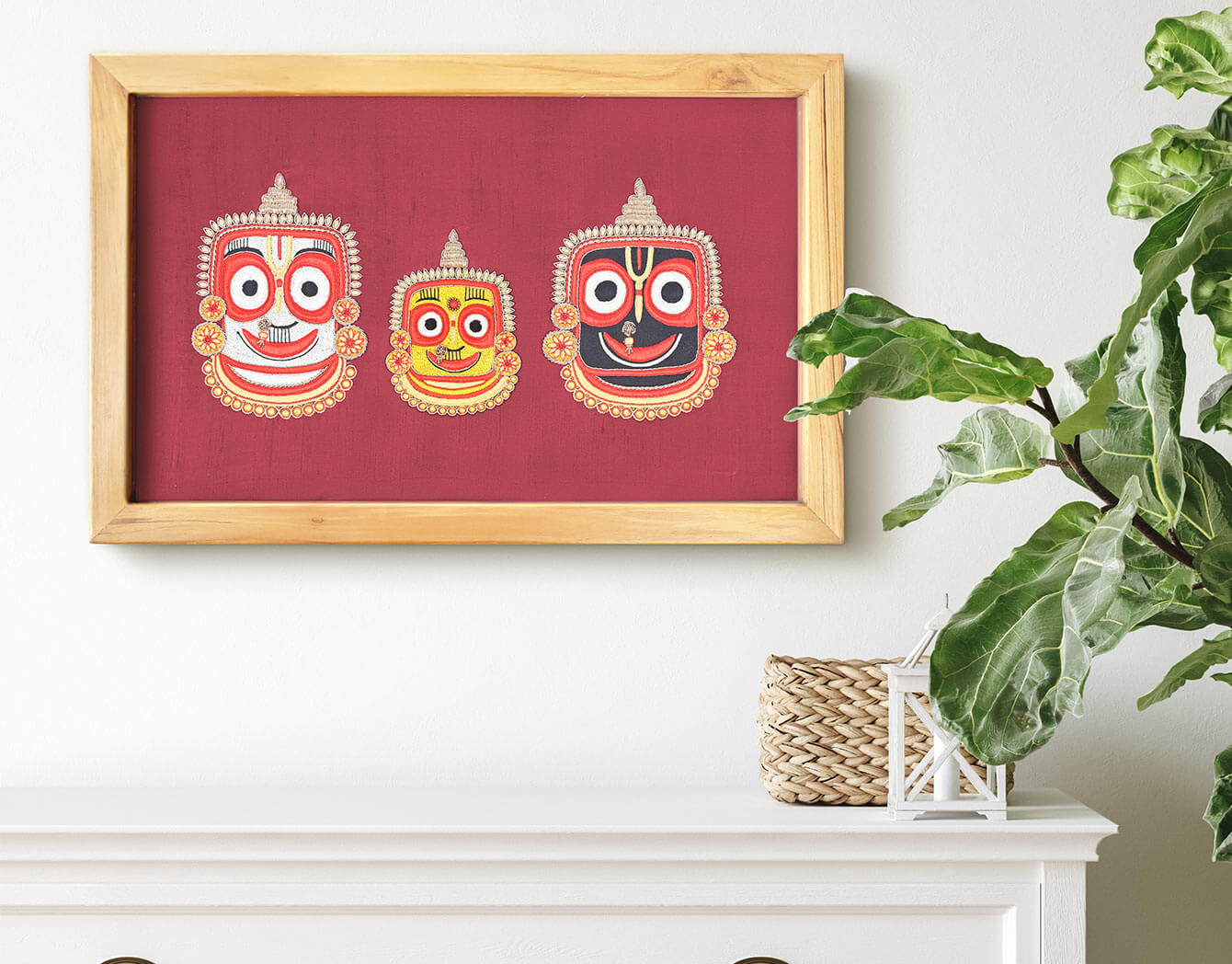Framed artwork with three colorful masks on a red background, placed on a white surface with a plant in the background.
