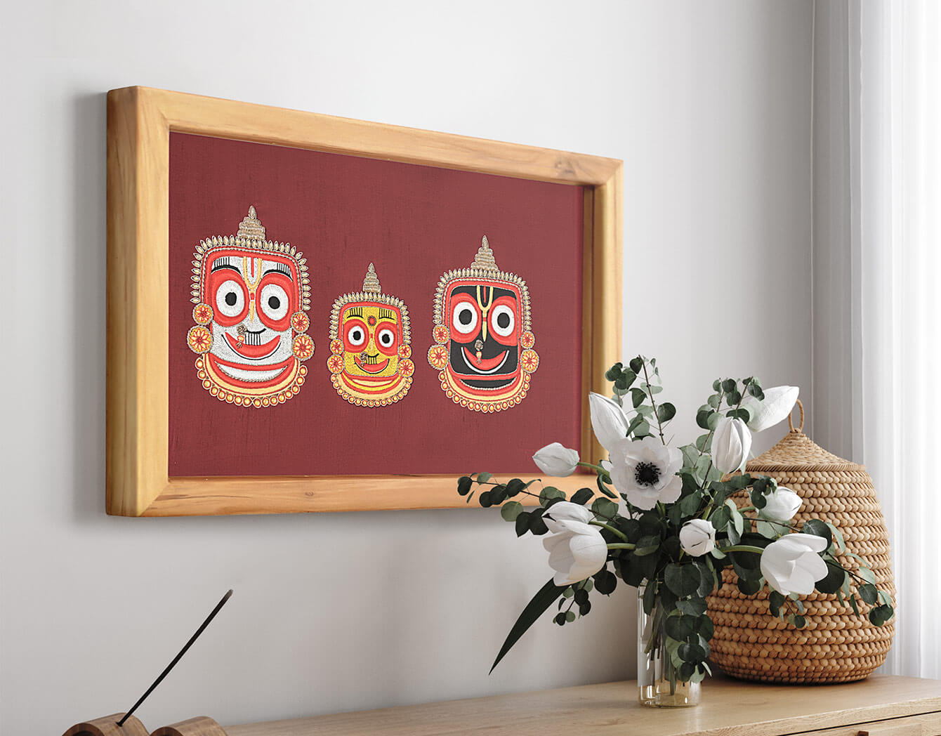 Framed artwork with two colorful masks on a red background, displayed on a wall above a wooden surface with a vase of flowers and woven basket.