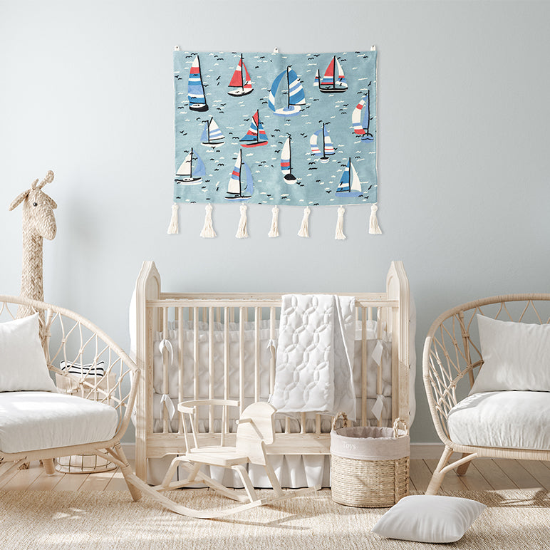 Hand-embroidered sailboat wall hanging of colorful boats on a blue sea—hung above a natural-wood crib in a light, airy nursery.