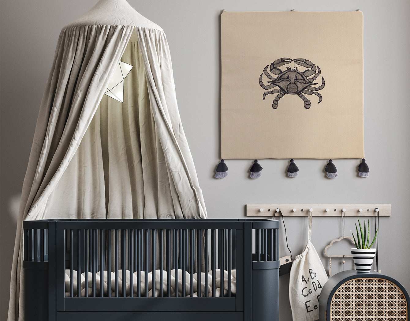 Embroidered textile wall hanging on taupe fabric with black tassels—styled above a grey crib in a modern nursery; coastal wall decor.