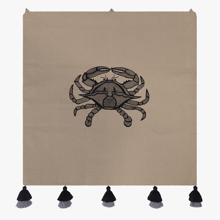 Embroidered wall hanging with a black crab motif on taupe fabric, finished with black-and-grey tassels along the bottom.