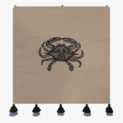 Embroidered wall hanging with a black crab motif on taupe fabric, finished with black-and-grey tassels along the bottom.