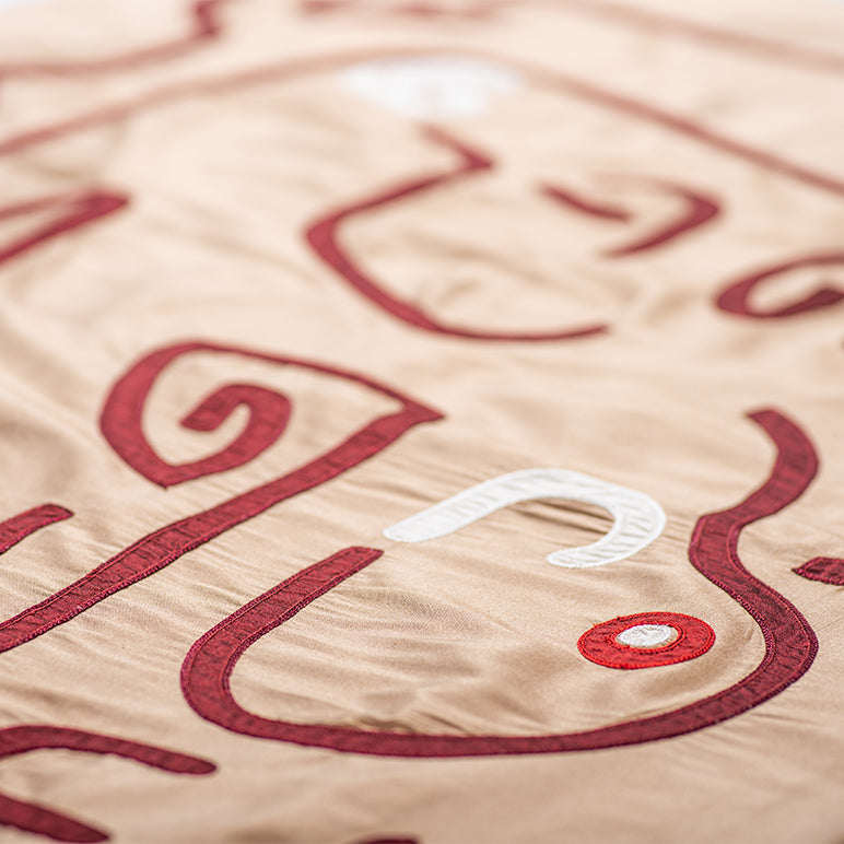 Close-up of the Crimson Whispers tapestry—raised crimson embroidery and a small white-and-red dot on warm beige fabric.