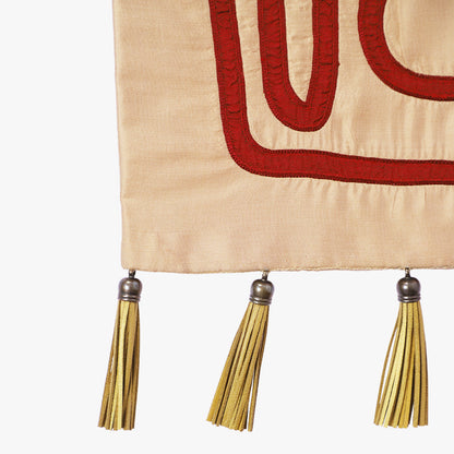 Close-up of the Crimson Whispers tapestry—crimson embroidered lines on warm beige fabric with gold tassels and metal caps along the bottom edge.