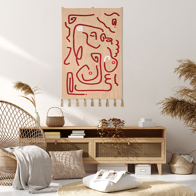 Crimson Whispers tapestry—abstract crimson line art on warm beige fabric with gold tassels—hung above a natural-wood console in a light, boho living room with woven accents.

