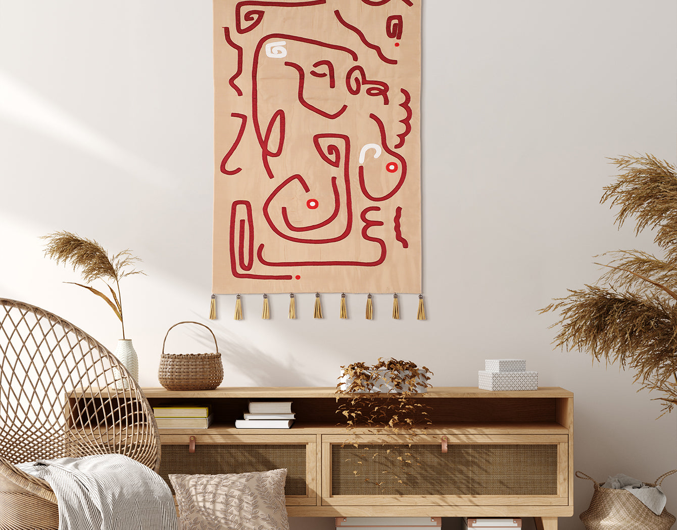 Crimson Whispers tapestry—abstract crimson line art on warm beige with gold tassels—styled above a natural-wood console in a light, boho living room with woven chair and dried botanicals.
