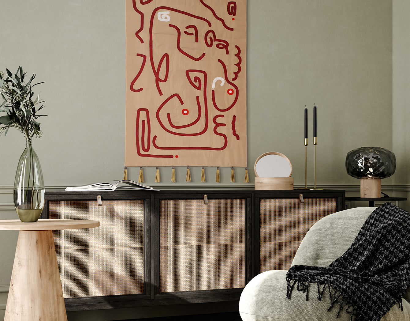 Crimson Whispers tapestry—abstract crimson line art on warm beige with gold tassels—hung above a dark wood cane console in a sage-green room with modern chair, black candlesticks, and a smoky glass lamp.