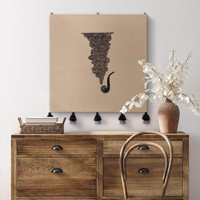 Wooden dresser with a vase of flowers and a chair, a decorative embroidered wall hanging above on a white wall.
