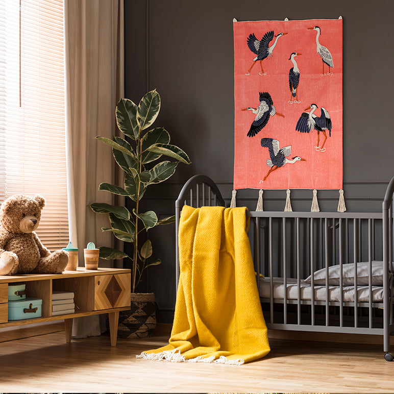 Embroidered wall hanging with navy-white herons on pink—hung above a grey crib with a yellow throw in a cozy kids’ room.