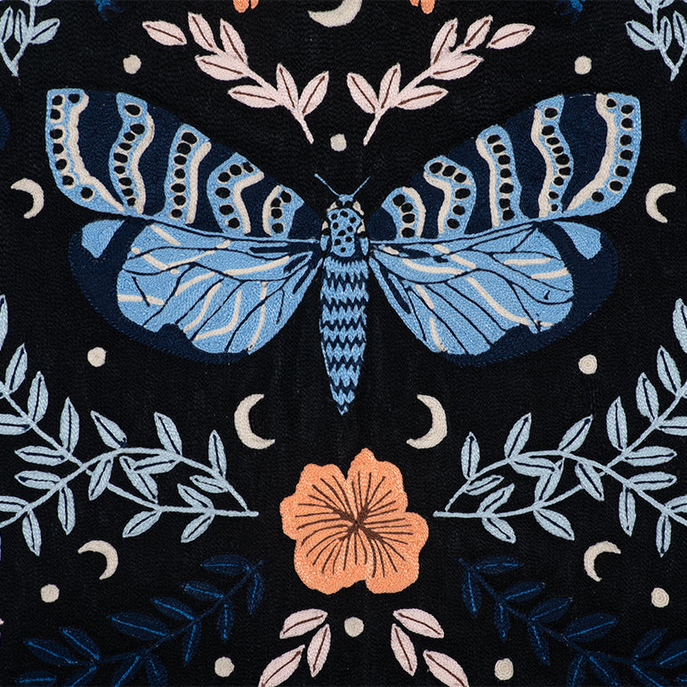 Close-up of Night Garden embroidered wall hanging—a blue moth with patterned wings surrounded by leafy vines, crescent moons, and a peach flower on black fabric.