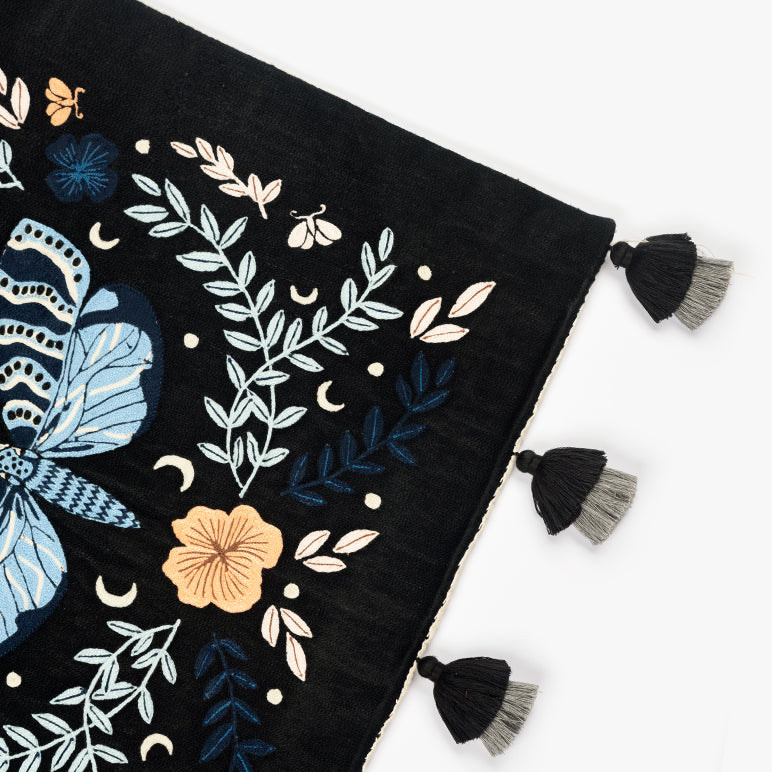 Corner detail of **Night Garden** embroidered wall hanging—blue moth and leafy vines with crescent moons on black fabric, finished with black-and-grey tassels.

