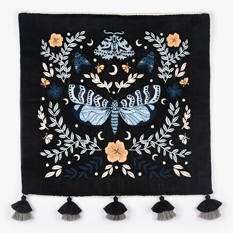 Night Garden embroidered wall hanging—moth and floral motif in blue and cream on black—hung above a grey crib with a yellow throw in a cozy nursery.
