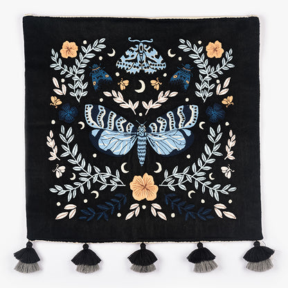 Night Garden embroidered wall hanging—moth and floral motif in blue and cream on black—hung above a grey crib with a yellow throw in a cozy nursery.
