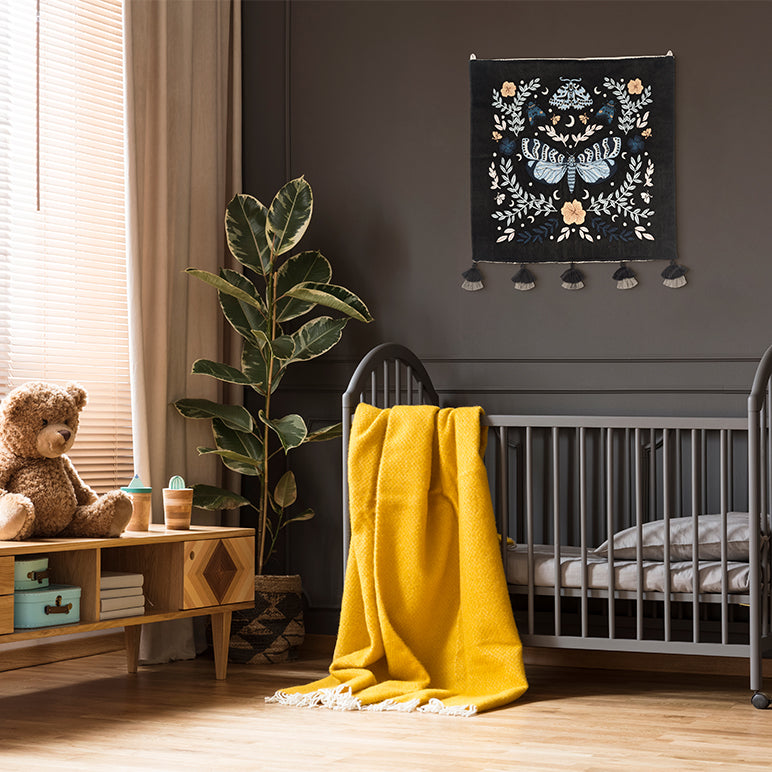 Night Garden embroidered wall hanging—moth and florals in soft blues and cream on black—hung above a grey crib with a yellow throw in a cozy nursery.
