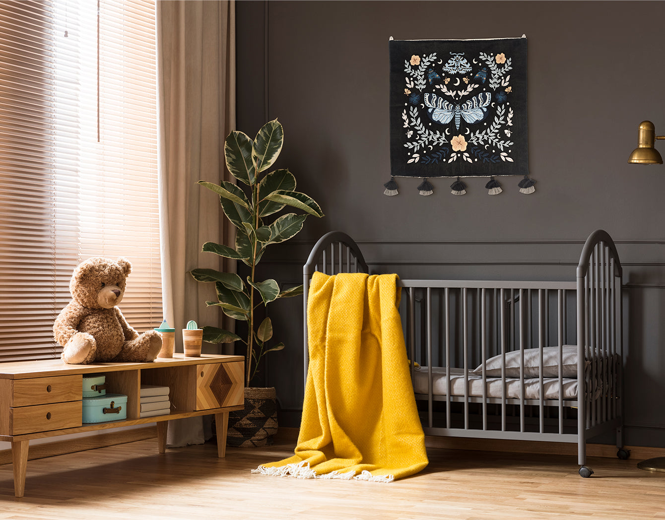 Night Garden embroidered wall hanging—blue moth and florals on black—hung above a grey crib with a bright yellow throw in a cozy nursery.