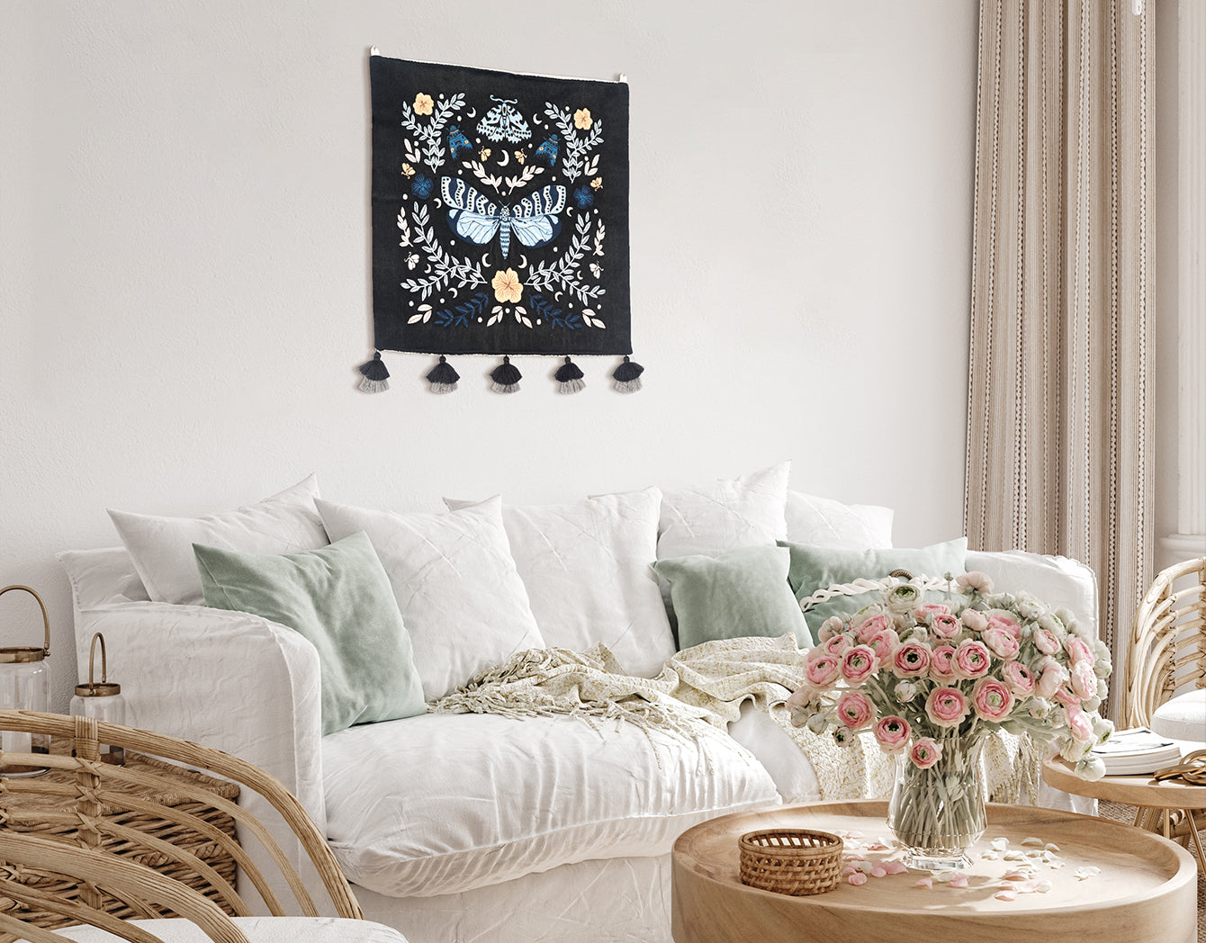 Night Garden embroidered wall hanging—blue moth and florals on black—displayed above a white sofa with sage cushions and a wood coffee table with pink flowers.