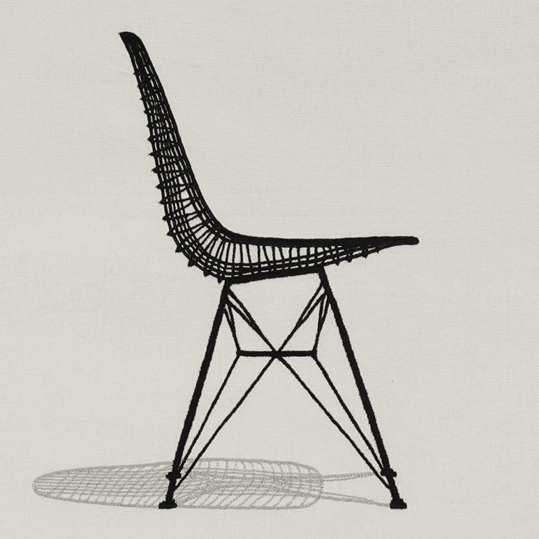 Close-up of the Sit Pretty embroidered wall hanging—black line-art chair stitched on off-white fabric with a grey shadow.