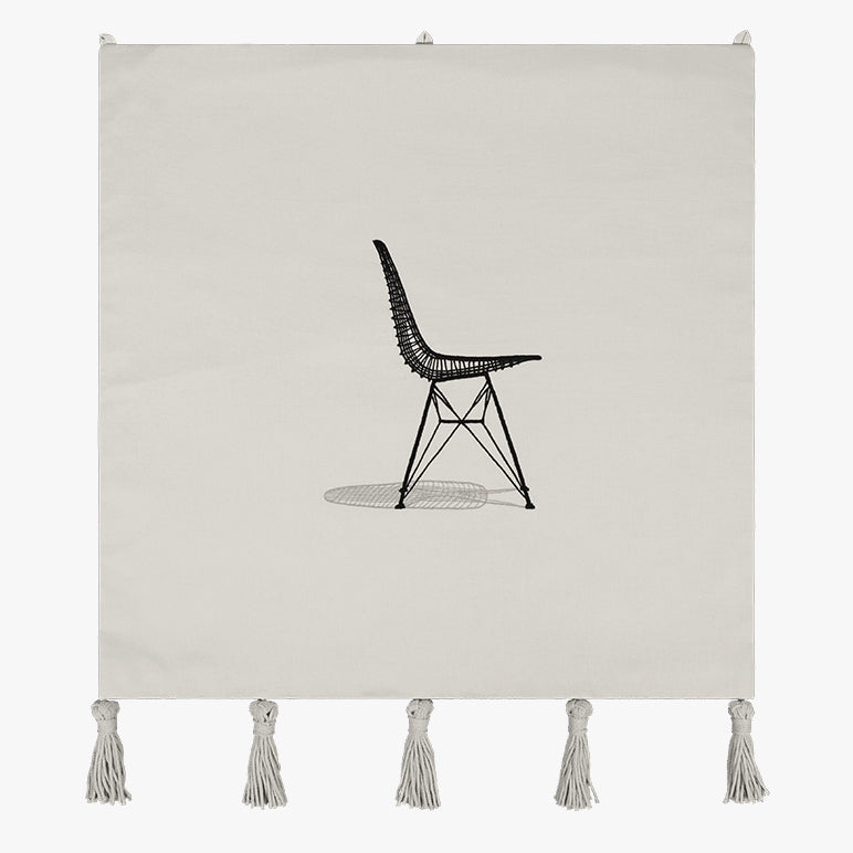 Sit Pretty embroidered wall hanging—minimal modern wall art of a black line-art chair on off-white textile with tassels; handmade textile wall art / tassel wall hanging.
