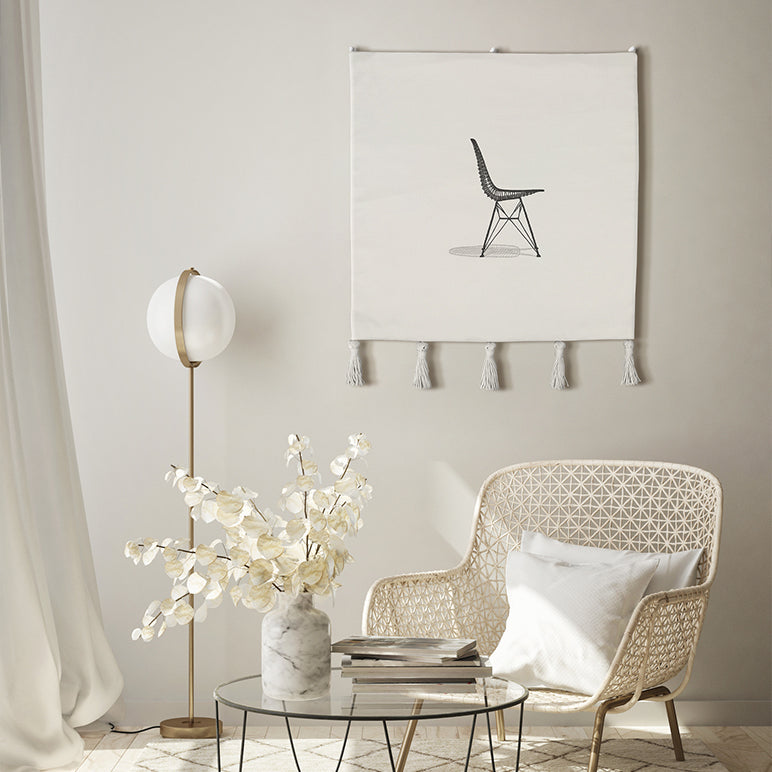 Embroidered wall hanging—minimal black chair on off-white textile with tassels—styled in a bright modern living room with cane chair and floor lamp; modern textile wall art.