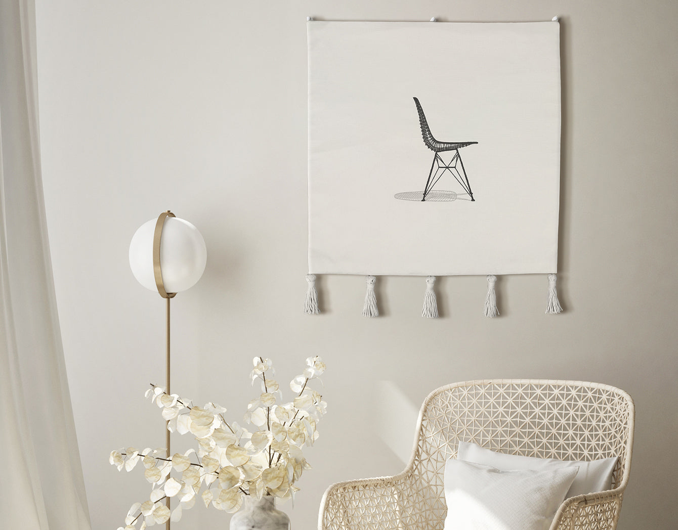 Sit Pretty embroidered wall hanging—black line-art chair on off-white textile with tassels—styled on a neutral wall beside a brass floor lamp and woven chair; modern minimalist living room.