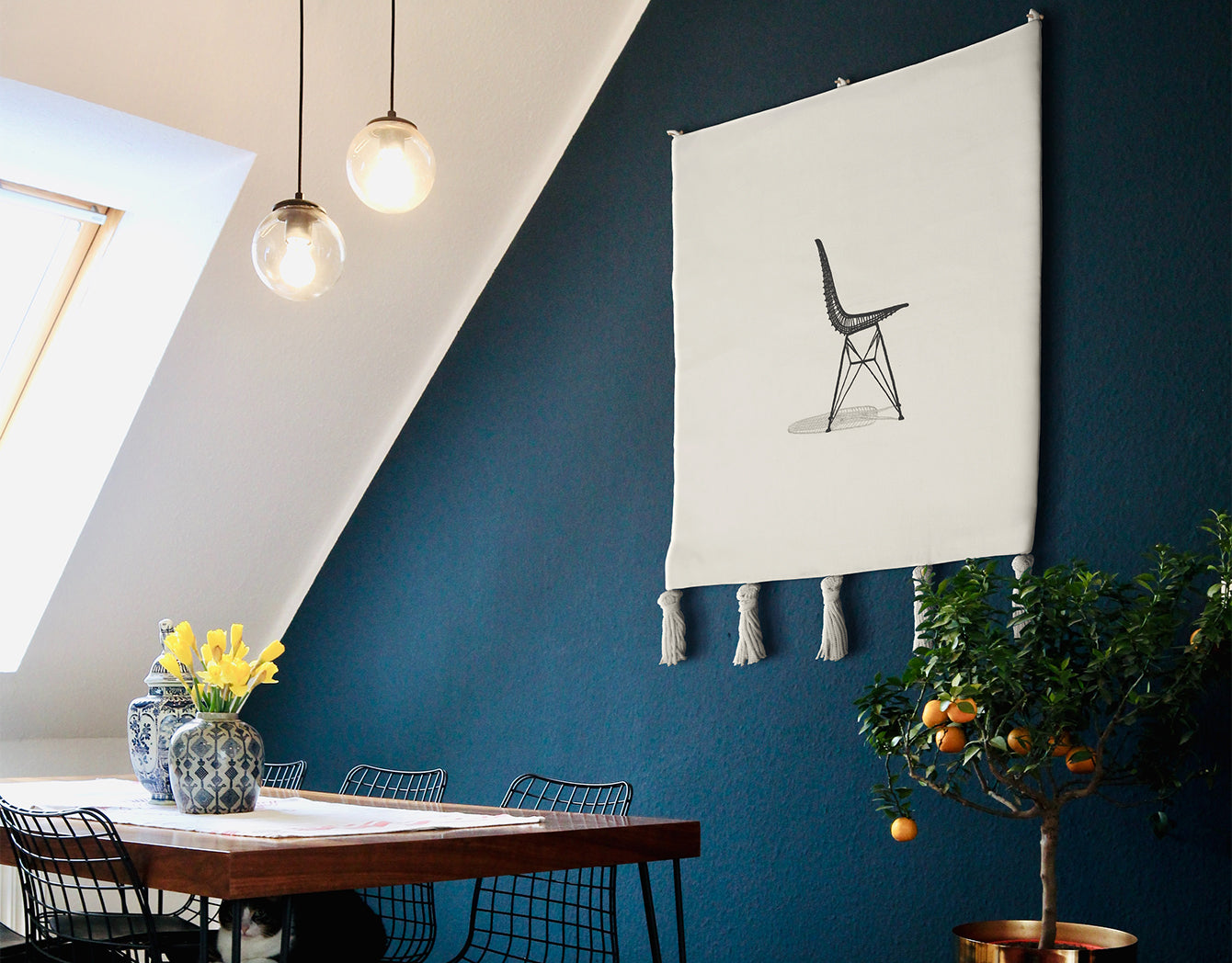 Sit Pretty embroidered wall hanging—black line-art chair on off-white textile with tassels—hung on a deep blue dining room wall above a wooden table with pendant lights; modern minimalist wall art.