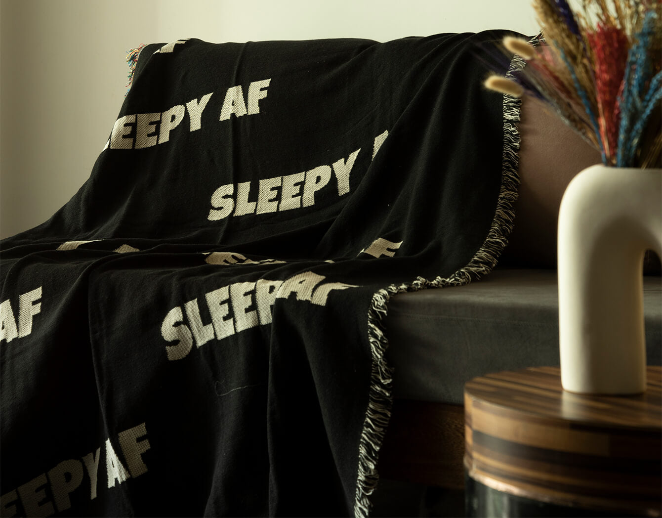 Black blanket with 'SLEEPY AF' text draped over a couch in a cozy living room setting.