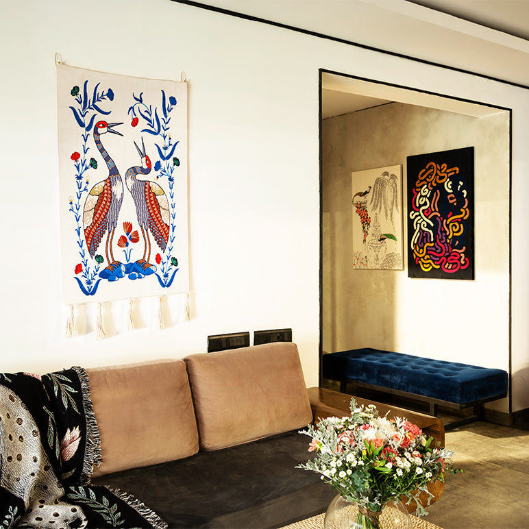 Songs of the Crane- textile wall art, a hand-embroidered wall hanging with two stylized cranes and blue floral vines, shown as modern wall decor above a tan sofa.