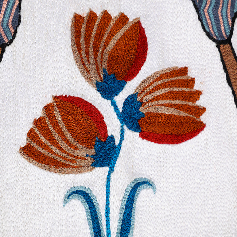 Close-up of *Songs of the Crane* embroidered wall hanging—stitched rust-and-red flowers with teal stems on an ivory textile.

