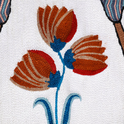 Close-up of *Songs of the Crane* embroidered wall hanging—stitched rust-and-red flowers with teal stems on an ivory textile.

