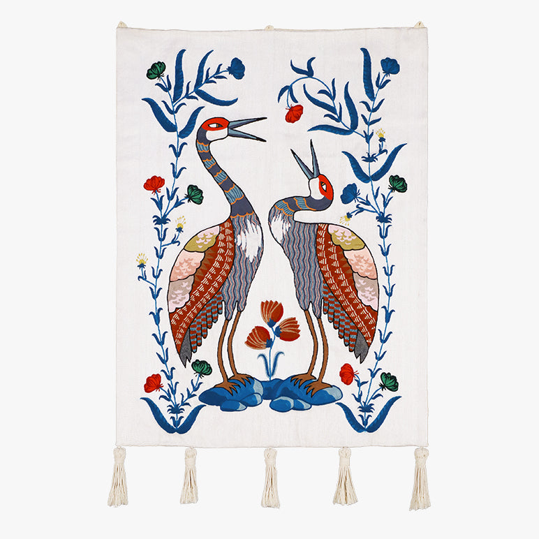 Songs of the Crane embroidered wall hanging—two stylized cranes with floral vines in blue, rust and green on an ivory textile, finished with tassels on a white background