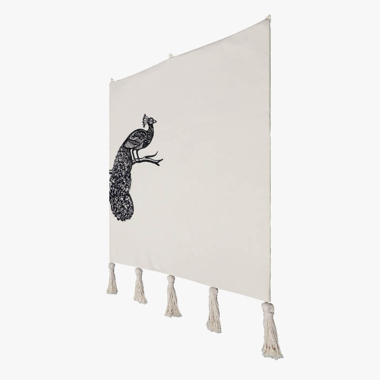 side angle view of a bird art tapestry with a hand-embroidered peacock on one side of the beige tapestry with macramé tassels