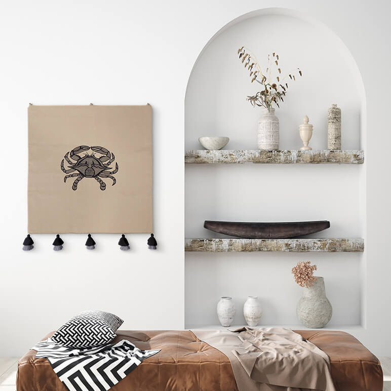 minimalistic crab wall art in the form of an embroidered tapestry with black tassels seen displayed in a modern white room with a brown leather bench