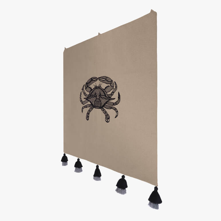 side view of a minimalistic crab art tapestry done in black thread work over an earthy toned cotton background, seen with five black tassels at the bottom