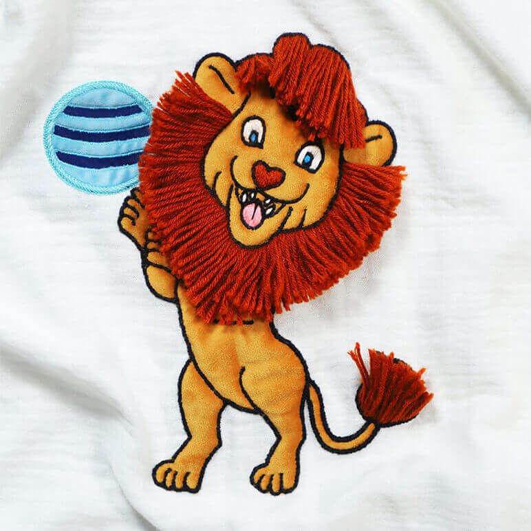 embroidered cartoonish lion in mustard and orange colors playing with a blue ball over a white bamboo cotton fabric background